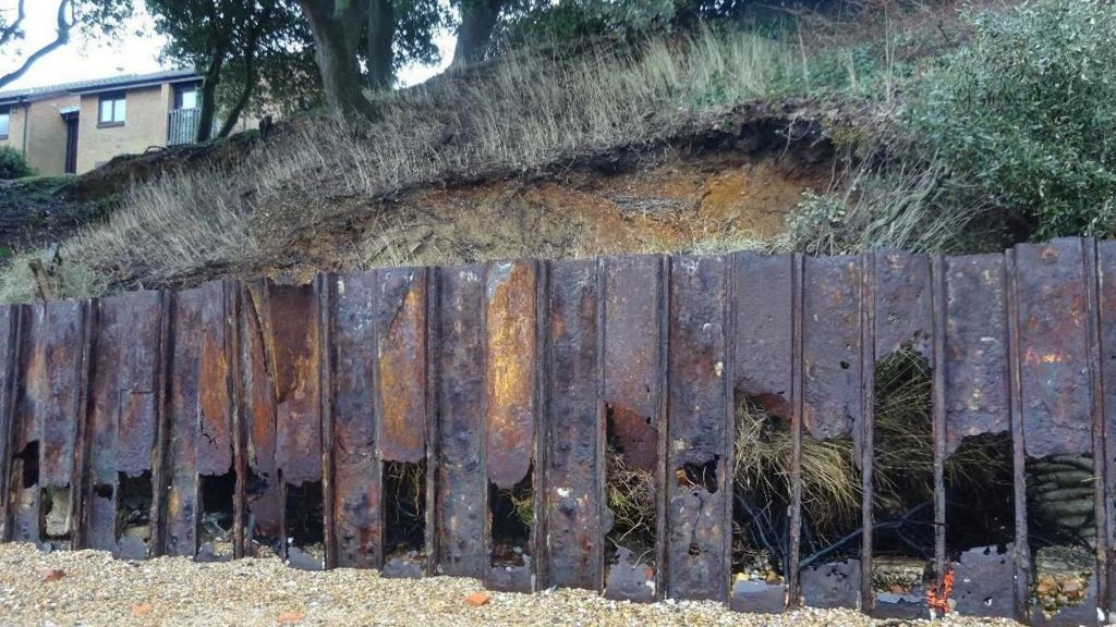 A severely rusted steel sheet pile wall at the bottom of a wooded slope leading to a stony beach. A private property can be seen on the top of the slope