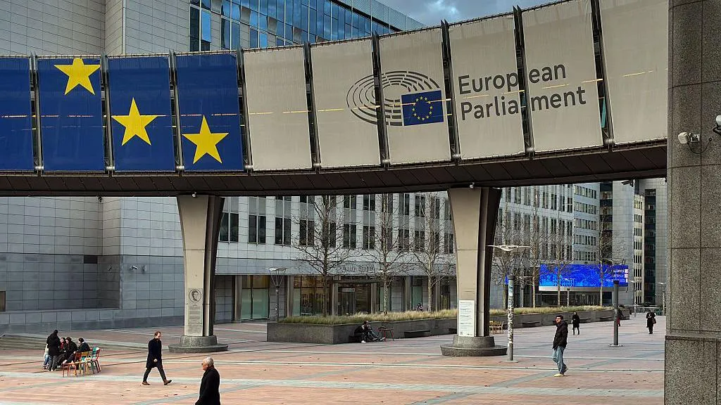 An exterior view of the Altiero Spinelli Building, part of the legislative complex of the European Parliament, shows a banner sign with the blue-and-yellow EU flag and the institution's name displayed on an elevated walkway.