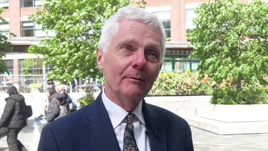 A man in a blue suit and patterned tie speaks in front of trees and white stone benches.