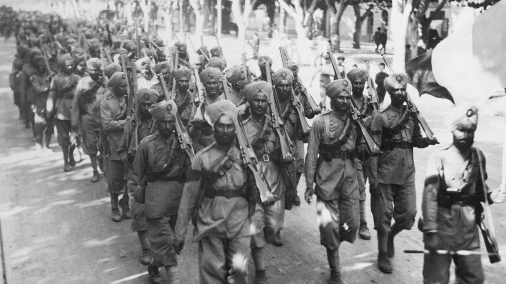A black and white image of a large group of Indian troops, wearing soldiers' uniforms, turbans, and holding guns, walk along a road