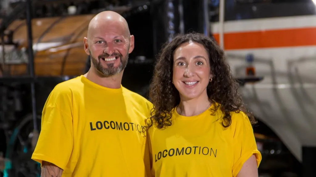 Two dancers, a bald man and a woman with brown curly hair, are smiling and wearing yellow tops with 'Locomotion' written on them.