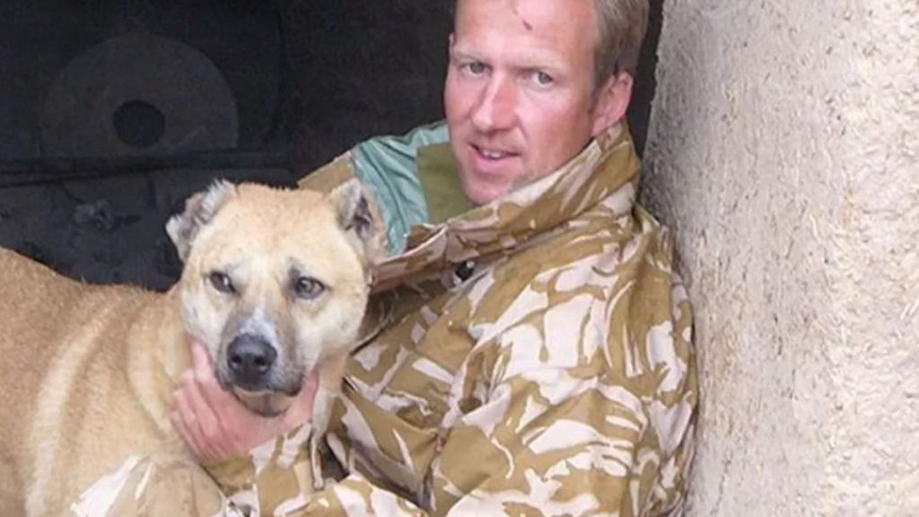 Pen Farthing looks directly at the camera as he holds a dog, he is sitting down and wearing Royal Marine commando gear. 