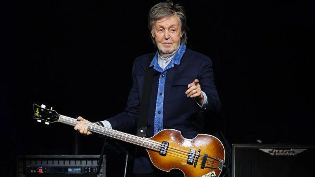 Sir Paul Mccartney stands on stage with a guitar and pointing towards the crowd