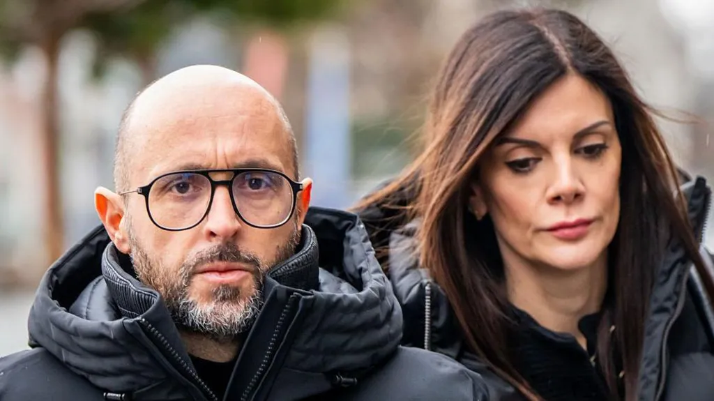 A bearded man with no hair and an anorak on the left arrives with a woman to his right with brown hair