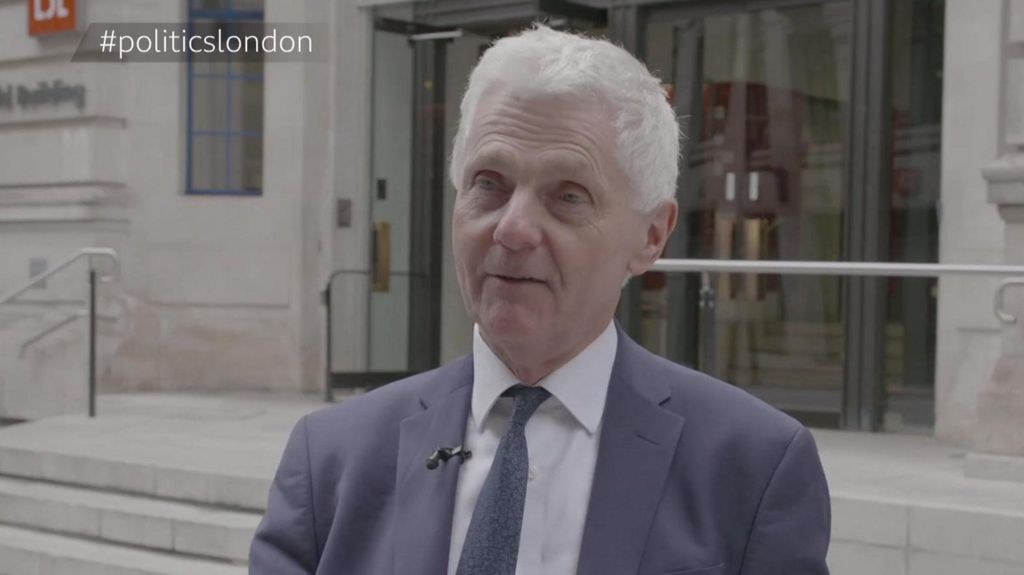 A middle aged man with white hair is being interviewed standing outside a civil building. He is wearing a grey/blue suit and matching tie and a white shirt. 