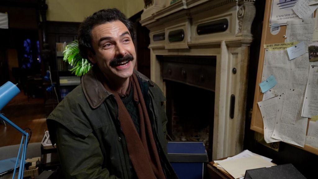 Aiden Turner laughs as he speaks to someone off camera. He's wearing a green jacket with brown scarf, and has a thick moustache.