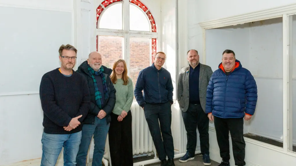 Six people - five men and one woman - stand in a row inside an empty room. The room is painted white and they are standing in front of a window. They are all smiling at the camera. 