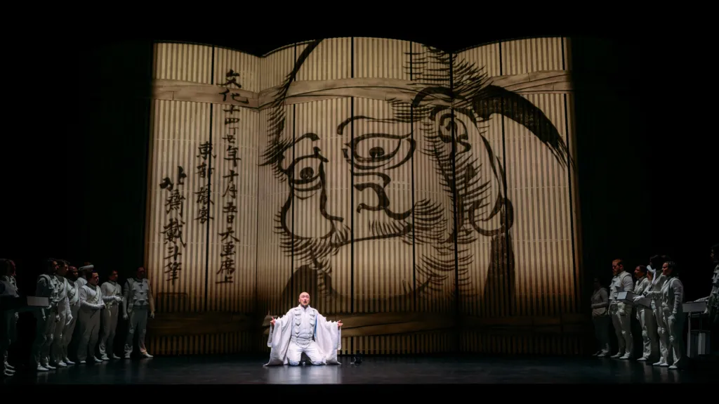 A man dressed in white kneels on the stage. A black ink drawing of a man with Japanese script is displayed in the background.