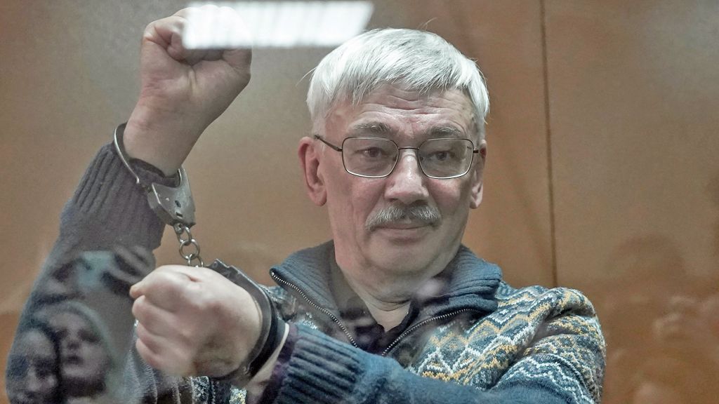 Oleg Orlov pictured behind the glass of a court dock, holding his arms in the air to show he is wearing handcuffs and clenching his first with a slight smile on his face. He wears a thick patterned jumper with a zip and collar, he has silver hair, a moustache and wire-rimmed glasses.