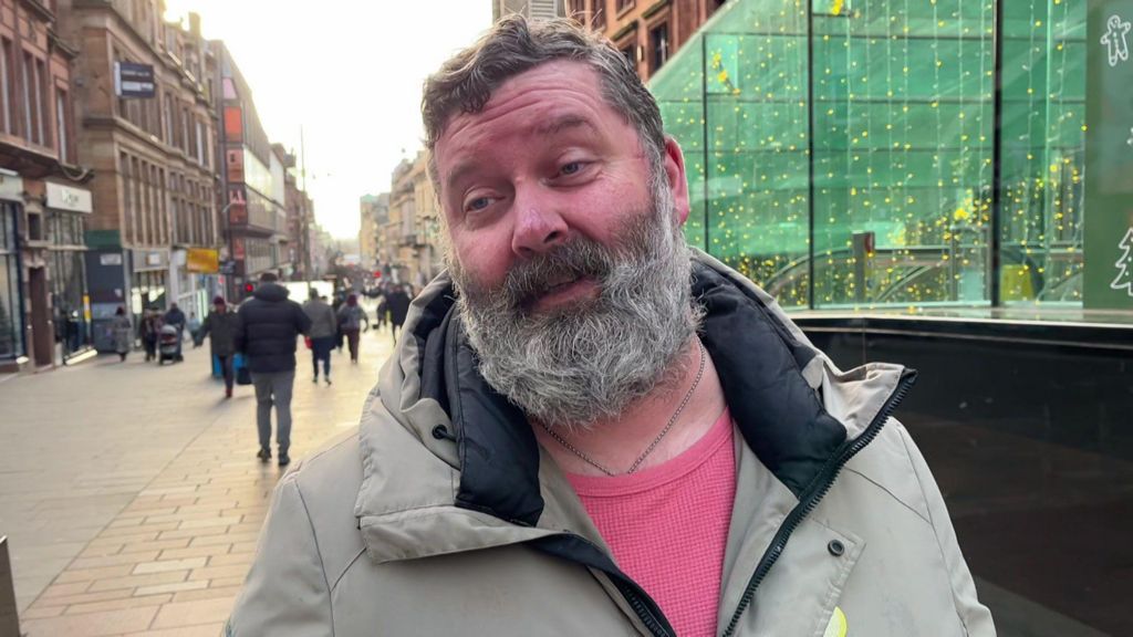 A man with a beard, smiling, wearing a necklace, and a pink jumper under a beige winter coat.