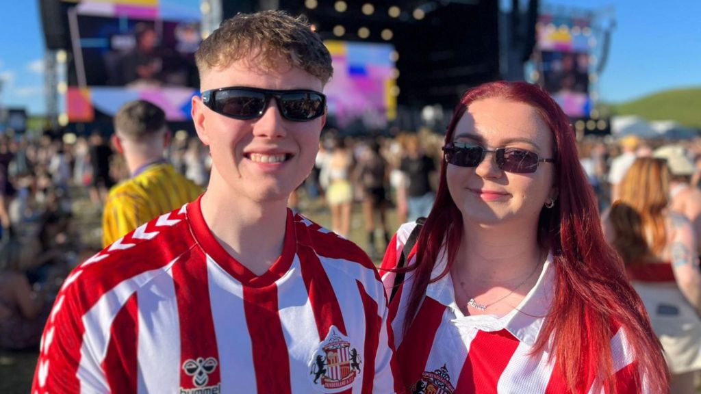 A young man and a young woman smile in front of the Big Weekend main stage and blue sky. They are both wearing red and white Sunderland football shirts and sunglasses. 