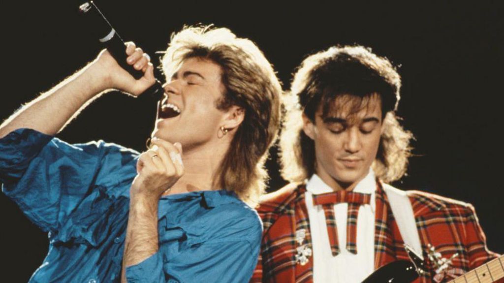 George Michael (left) wears a blue shirt and sings into a microphone he is holding up to his mouth, while Andrew Ridgeley looks down at a guitar he is playing out-of-frame wearing a tartan suit, as they perform together live on stage in 1985.