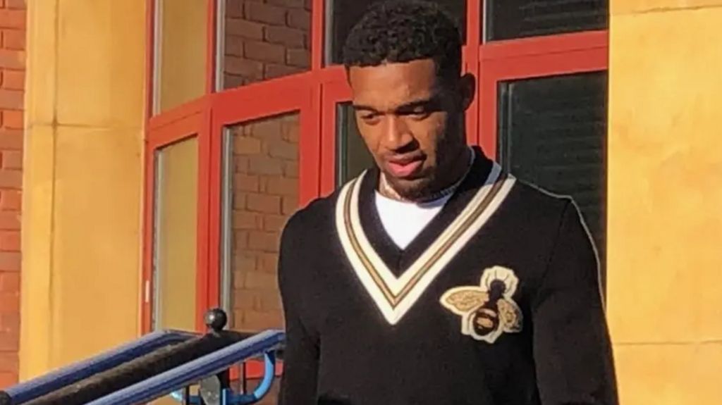 Jordon Ibe outside a building. He is wearing a black vest with a bee sewing on. He is looking down.