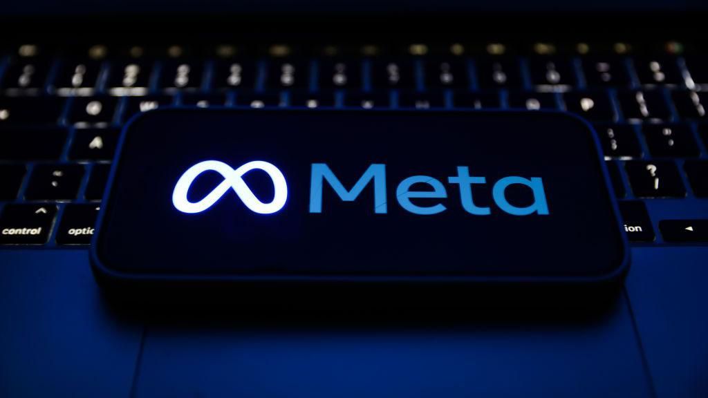 Smartphone displaying Meta's logo in blue, glowing against a black background. The phone is lying on a lit-up laptop keyboard. 