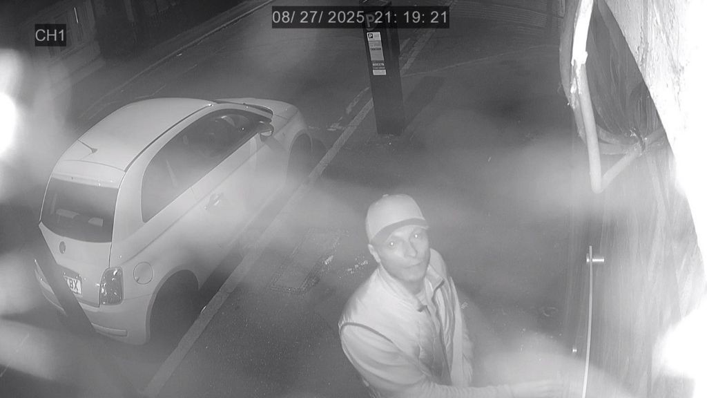 Black and white image of Muzyka looking up into the camera at a door. He is wearing a baseball cap and light-coloured clothes. A small parked car is also visible behind him.