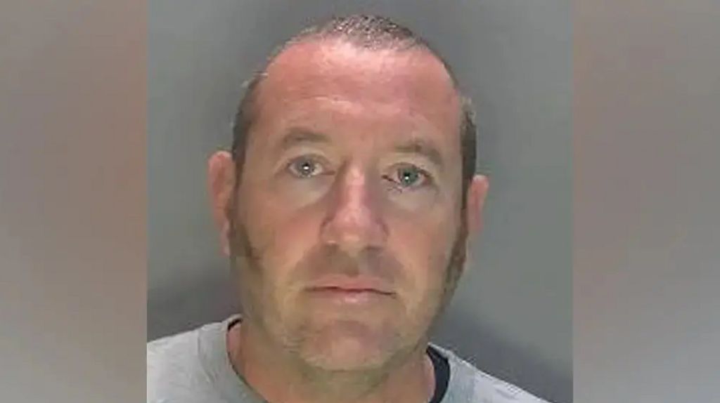 David Carrick photographed in police custody. He is wearing a grey sweatshirt and has sideburns and a near-shaven head. 