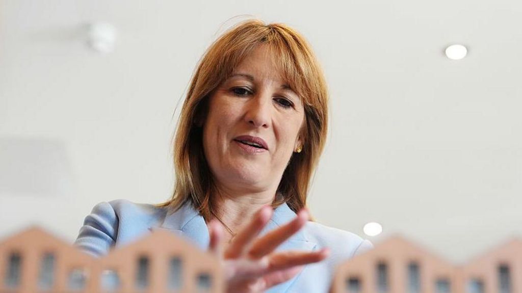 Chancellor Rachel Reeves, wearing a powder blue suit, standing with her hand over some model houses which are just seen in front of her