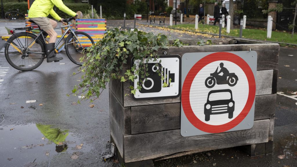 LTN box with a plant, camera sign and a red symbol with car and motorbike inside in the foreground. Behind, a cyclist goes past