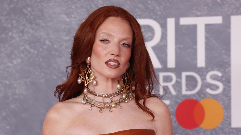 Jess is wearing a strapless dark orange dress. She has on large gold and pearl dangly earrings. She has long ginger hair past her shoulders and blue eyes. 