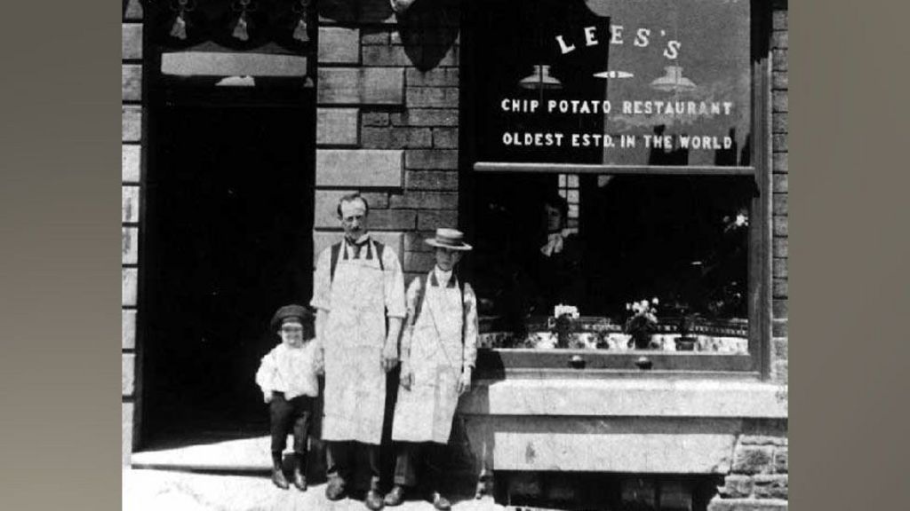 Black and white photo showing a child, man and boy standing in front of Lee's fish and chip shop
