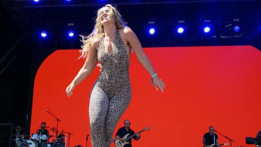 A woman in a leopard print leotard dancing on a big stage with a red bakckground and men playing instruments behind her