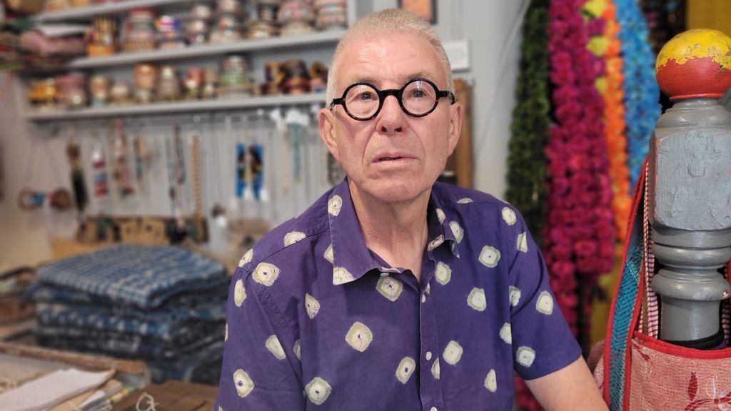 A man wearing a purple shirt and black glasses is sitting by a till in a shop. There is pottery and blankets behind him.