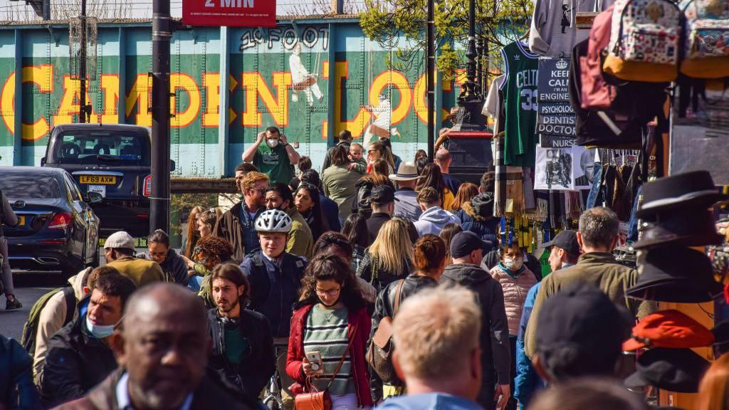 Image from 2021 showing crowds walking down Camden High Street, with the green and yellow Camden Lock bridge graffiti behind them