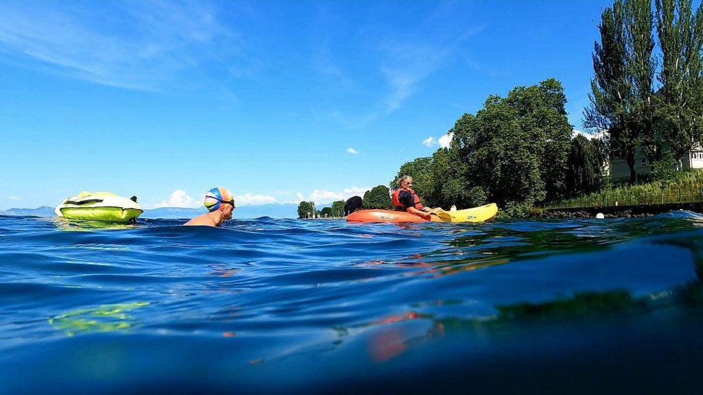 Ms Barratt is swimming in open water. She is wearing a multi-coloured swimming cap and dark goggles. She has a fluorescent yellow floatation device behind her. In front of her is a woman sitting in a yellow and orange kayak. On the edge of the water, there are dark green trees.