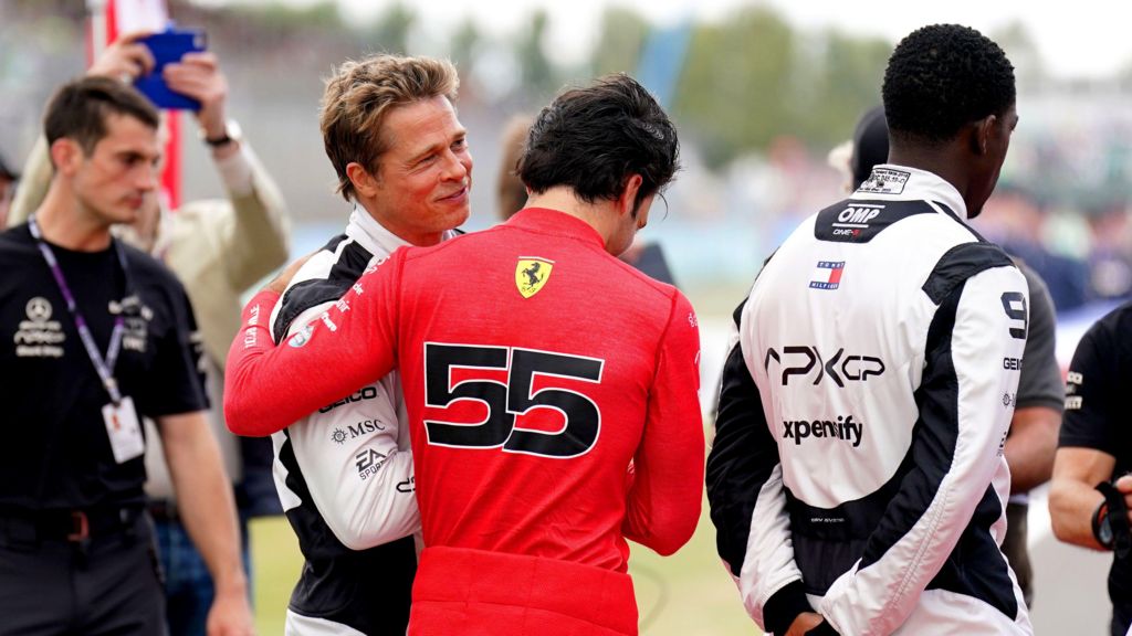 F1 driver Carlos Sainz, in a red/orange racing suit, embraces Brad Pitt, who is wearing a white racing suit.