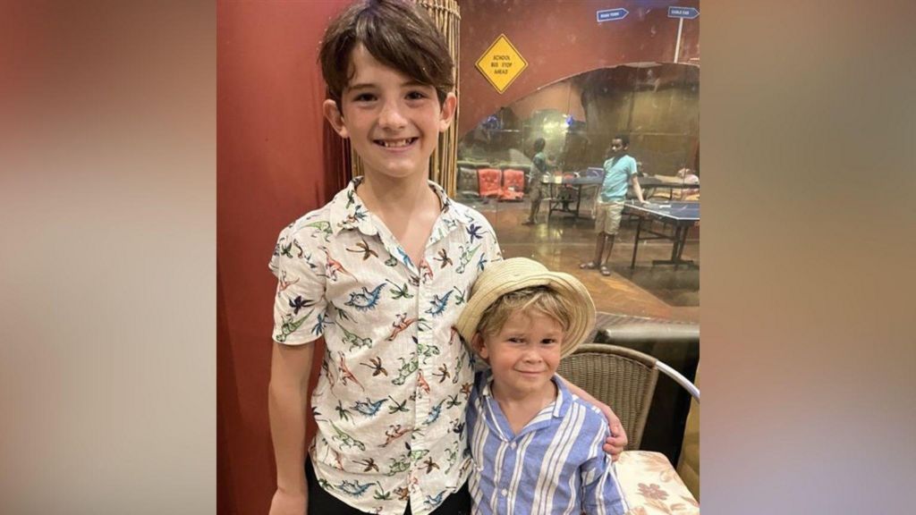 Rafael smiles at the camera as he stands with his arm around friend Fred Jones. Rafael has long brown hair and wears a white shirt with animals on it. Fred has a cream-coloured Panama hat on and a blue and white striped shirt. 