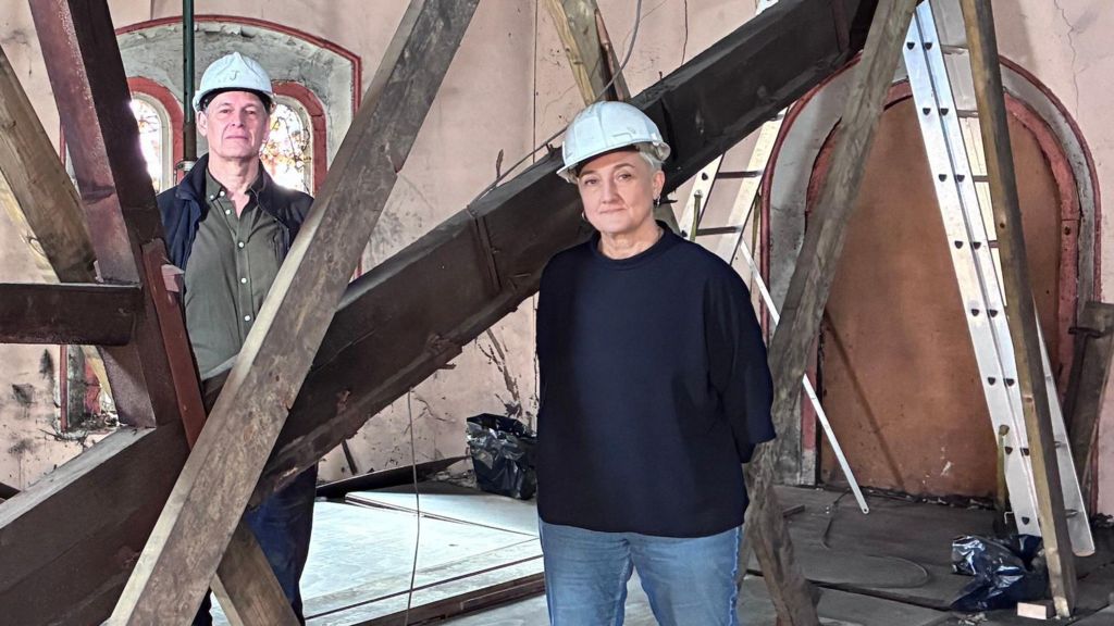 A man and a woman wearing white hard hats stand either side of a timber roof strut resting at an odd angle on the floor