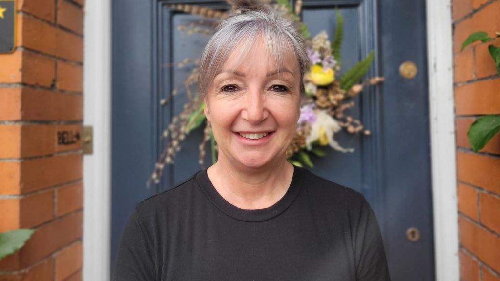 A woman in a black shirt is standing in front of a blue front door. She is smiling.