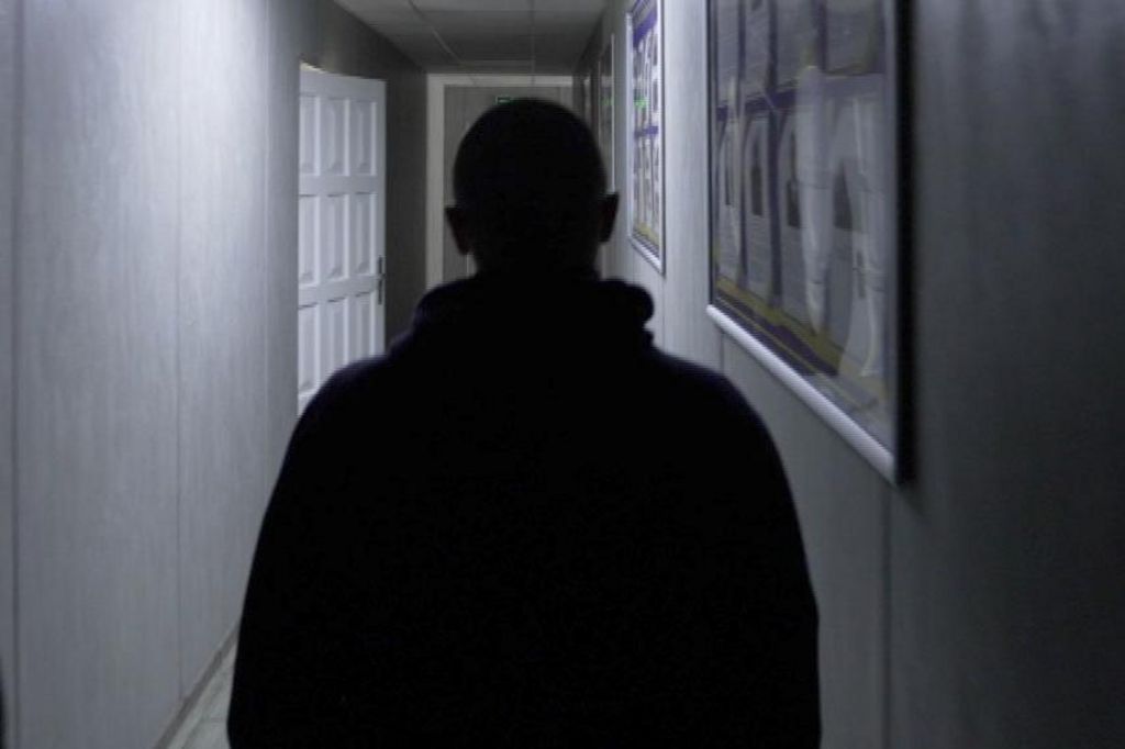 A rear view of the top half of Vlad in silhouette in a poorly-lit corridor of the Rivne detention centre. His head is shaven.