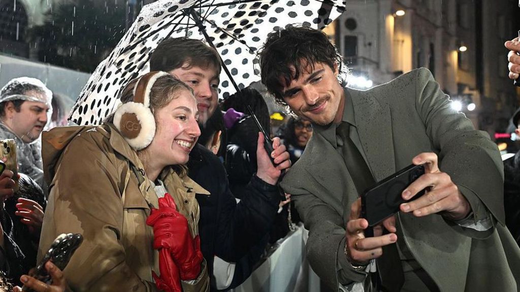 Jacob Elordi holds a phone up to take a selfie with two fans, one of whom is holding an umbrella, at the Wuthering Heights premiere. Elordi has dark hair and a moustache, and is wearing a grey suit.