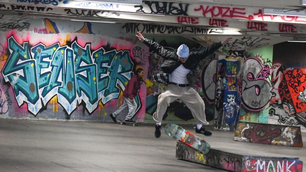Library photo of a skateboarder mid-jump. The background wall has graffiti tags on and it looks like it is in a similar building to a carpark