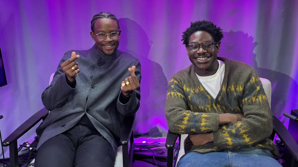 Two men sit on chairs in a studio with a purple curtain in the background. One man is dressed in a dark grey shirt, and smiles as he appears to click his fingers. The other man, who wears an olive cardigan with flashes of blue and yellow  pattern, smiles widely with his arms folded in a relaxed posture.