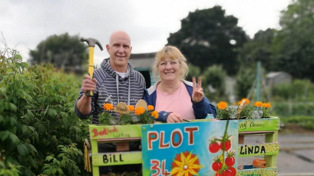 William in striped top holding hammer and Linda in pink top and blue hacket standing in an allotment