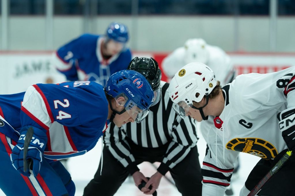 Two men from rival teams face each other on an ice hockey rink. On the left the man is in a blue and on the right the man is dressed in white. There is a referee in a black and white striped uniform in between them