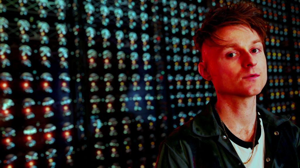 Look Mum No Computer stands in front of his 1,000 Oscillator Drone Synth, bathed in red light