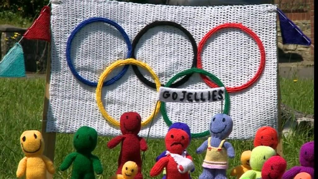 A group of multi-coloured, knitted figures stand in front of the Olympic logo. 