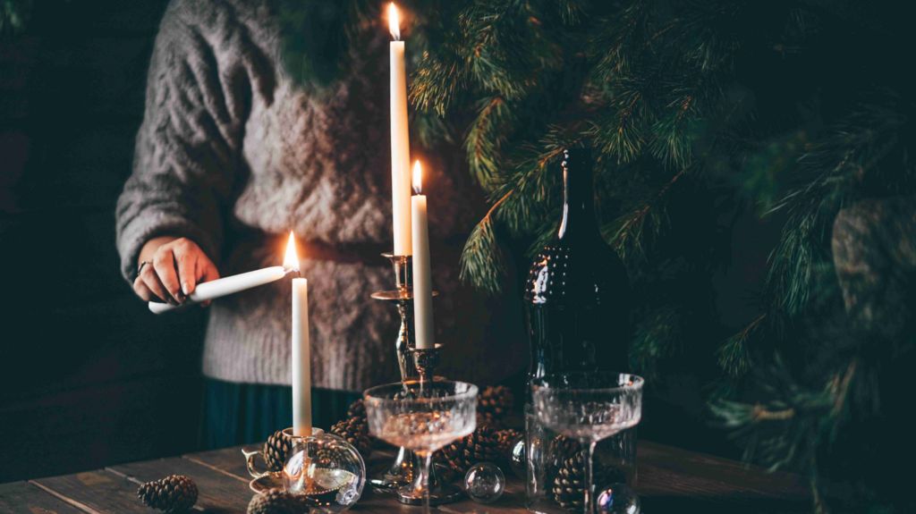 An unrecognizable woman in a cozy sweater lights tall candles on a rustic wooden table set for a quiet Christmas celebration. The warm candlelight, evergreen branches, and vintage champagne glasses create an intimate, hygge-inspired winter moment.