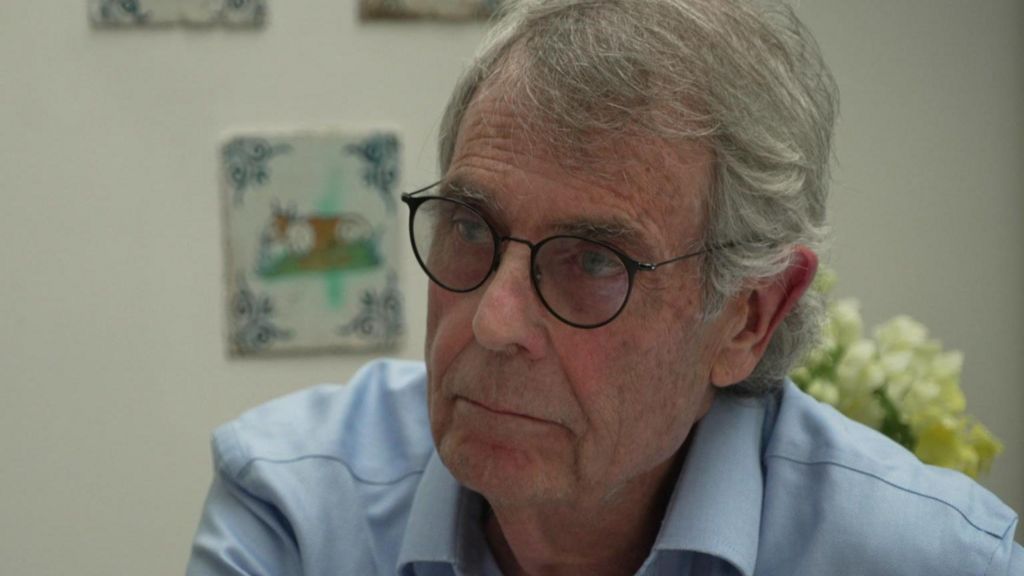 Peter Rook wears a pair of glasses and a light blue shirt. He is seated, in front of a wall with some artwork on it and flowers in the background.