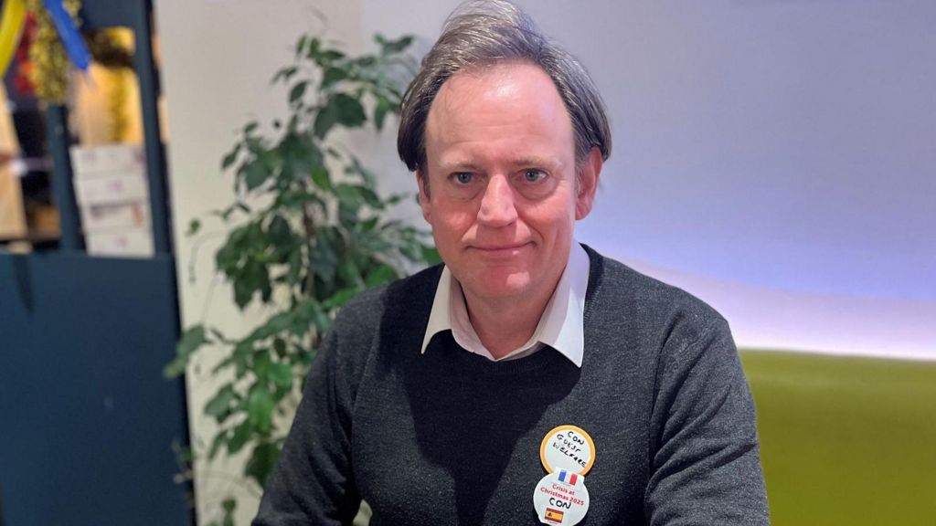 Con - a man with brown, floppy hair in his 40s or 50s. He wears a grey sweater and sits at a table with a large plant in the background.