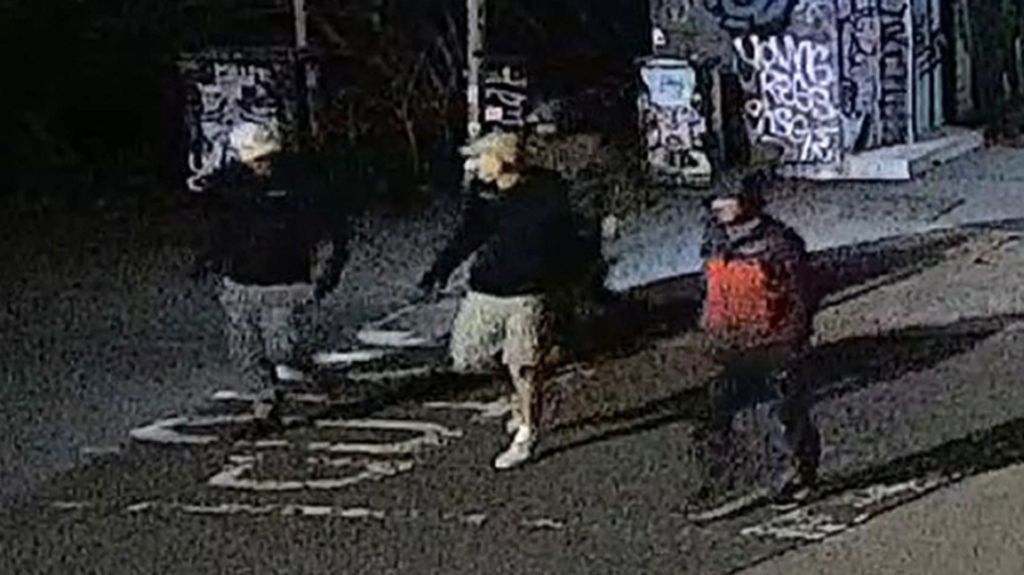 A blurry CCTV image of four men wearing jackets and baseball caps in a street at night time. 