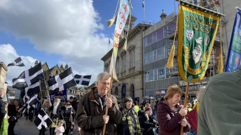 A crowd of people walking through Truro holding Cornish flags, wearing tartan scarves and black and white clothing.