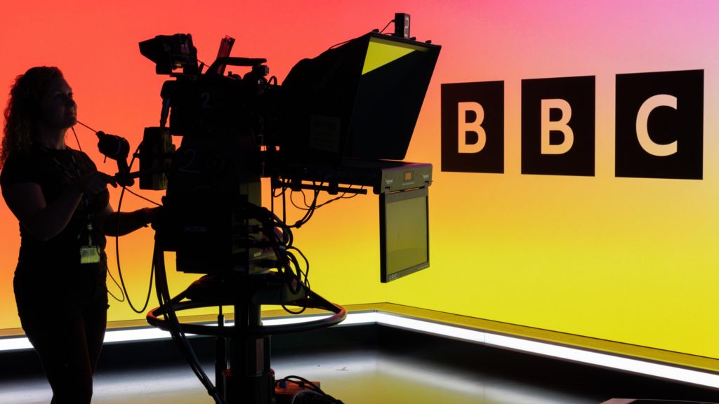 A woman operating a TV camera in front of the BBC Logo on an LED wall.