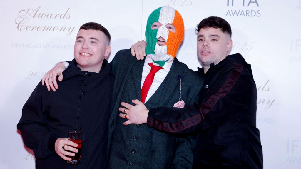 Members of Kneecap walk the red carpet at the IFTA Awards in Dublin. Mo Chara and Móglaí Bap are at either side of DJ Próvaí. They are wearing black and DJ Próvaí is wearing a green suit and a red tie. He is also wearing an Irish tricolour balaclava.