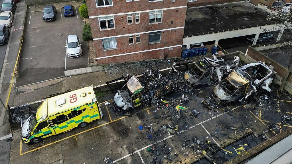 An aerial image shows burnt out ambulances in a parking area. Three vehicles at to the right are almost completely destroyed. There are cars and the lower levels of a block of flats visible at the rear of the frame.