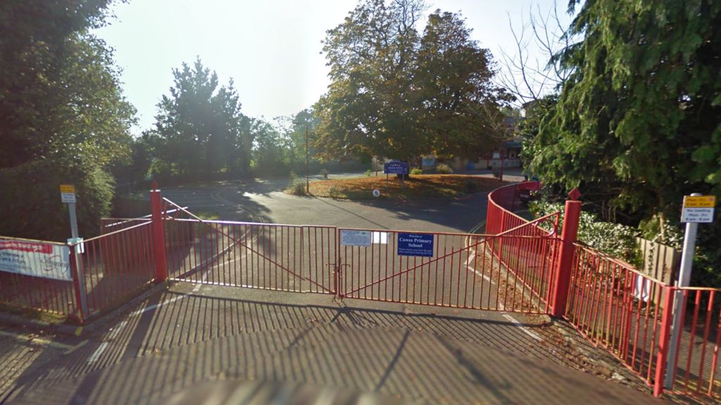 A screenshot from Google maps showing the outside of a primary school on a sunny day. It's got a red gate, which is closed, and a car park with a grassy island and a tree in the middle of it.