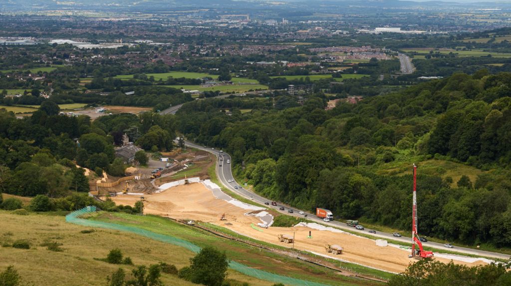 A road curves around next to a construction site. There are fields, a wooded area and homes in the distance.